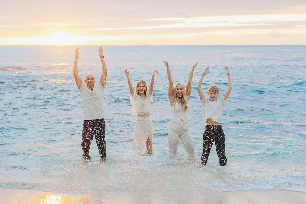 People jumping and splashing in crystal clear ocean water at sunset with pink sky reflections on the water's surface.