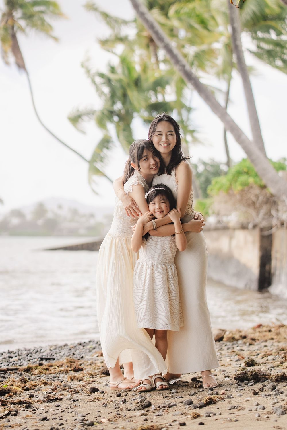A family wearing cream colored dresses poses together on a rocky beach with palm trees in the background.