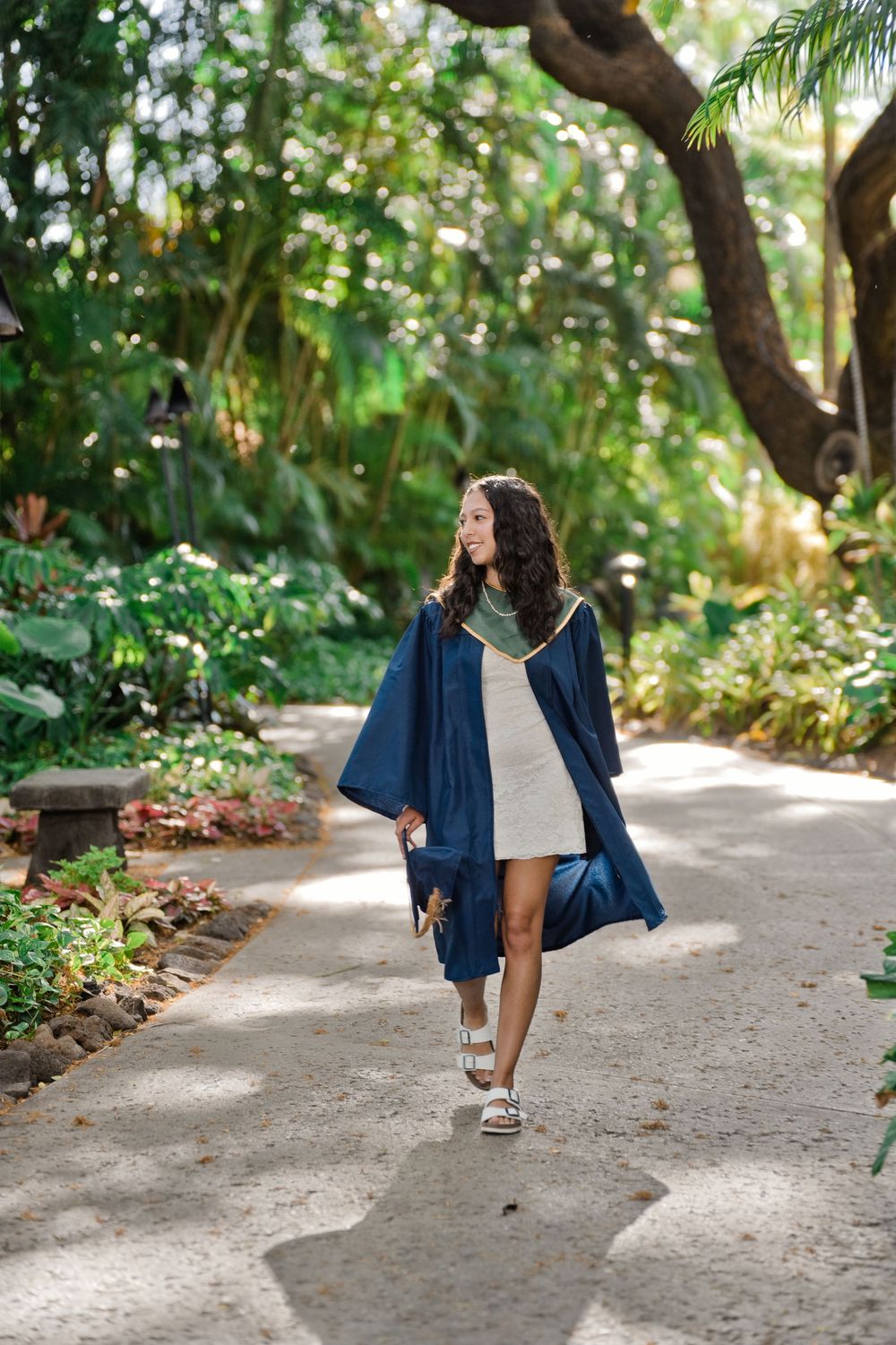Young woman in blue graduation gown walks along a lush tropical garden path, smiling and holding her cap.