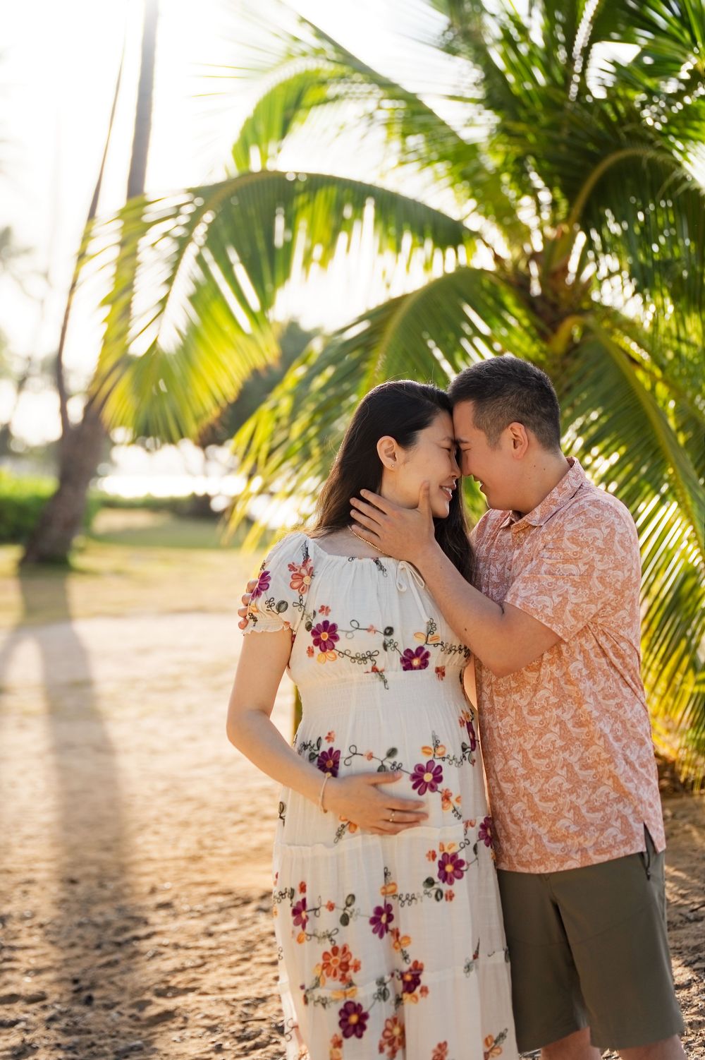 Couple sharing a tender moment on a tropical beach, pregnant woman in floral dress with partner under palm trees at sunset.