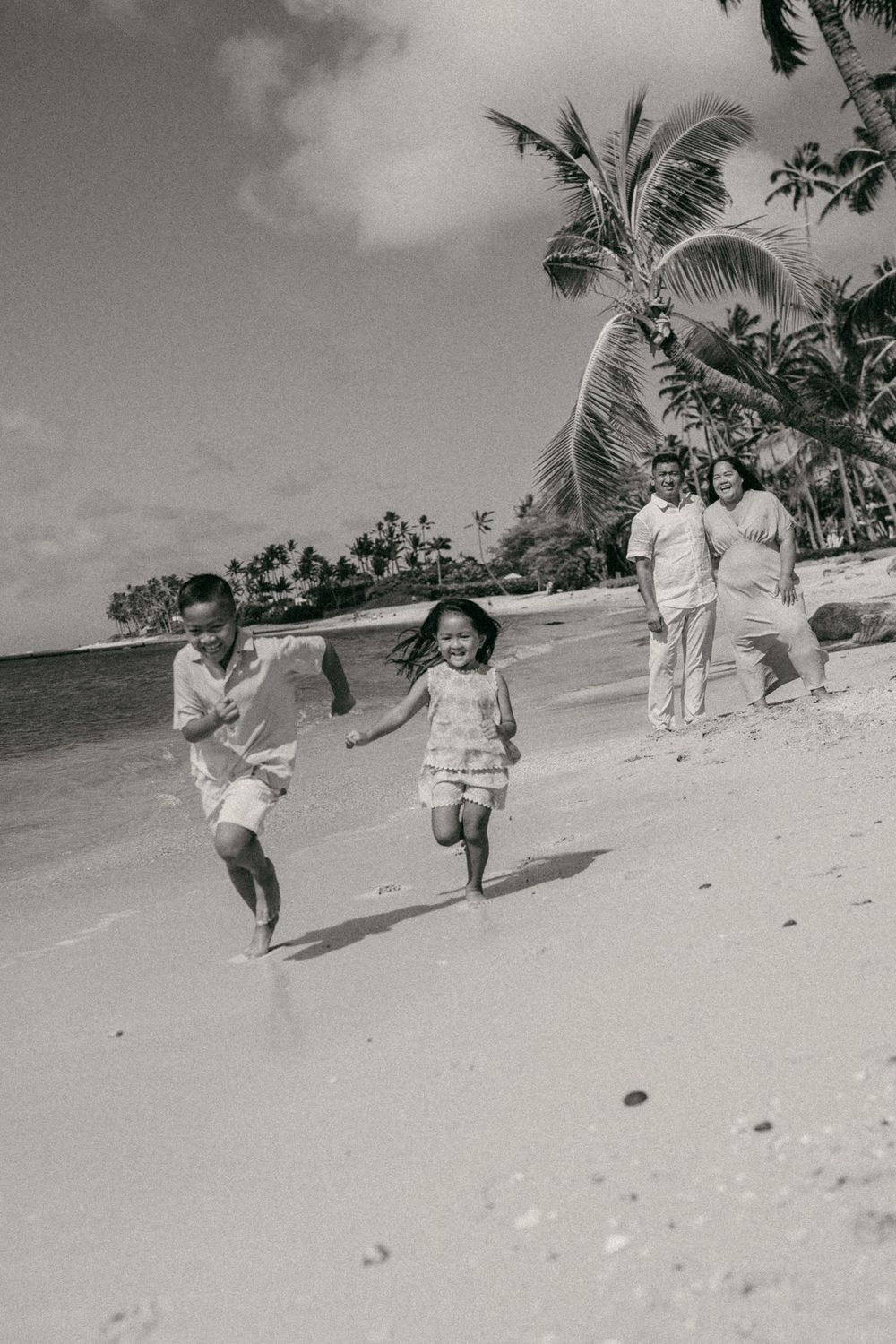 Black and white photo of two children running on a tropical beach while parents watch from behind near palm trees.