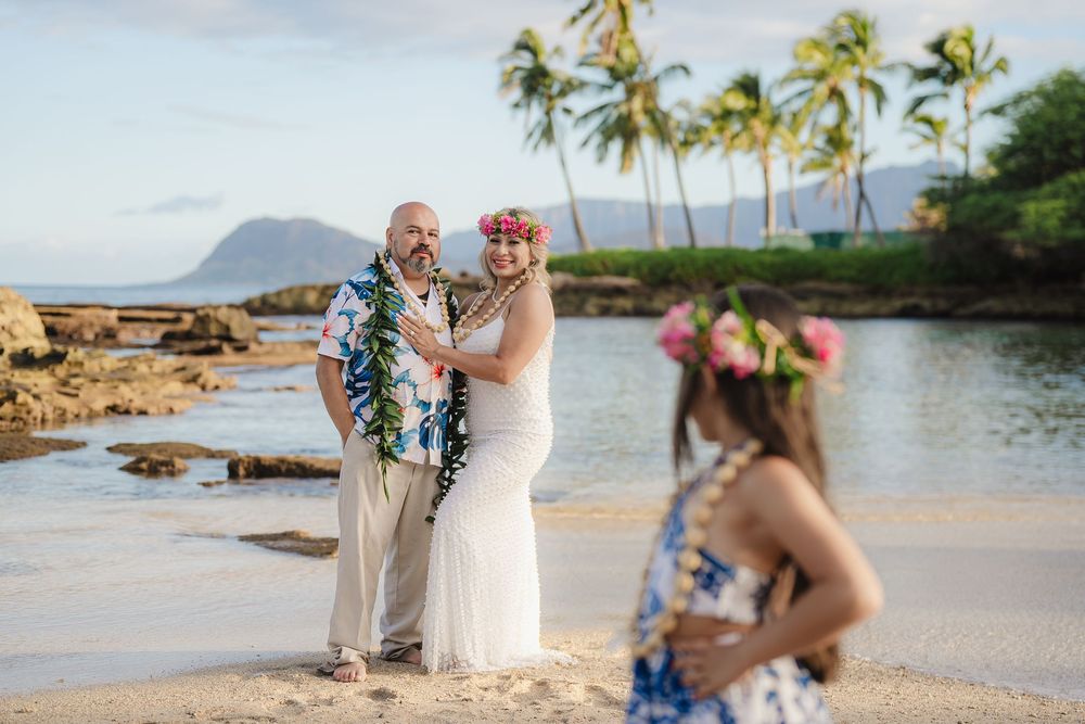 Couple in wedding attire with leis on a Hawaiian beach, with palm trees and mountains in the background.
