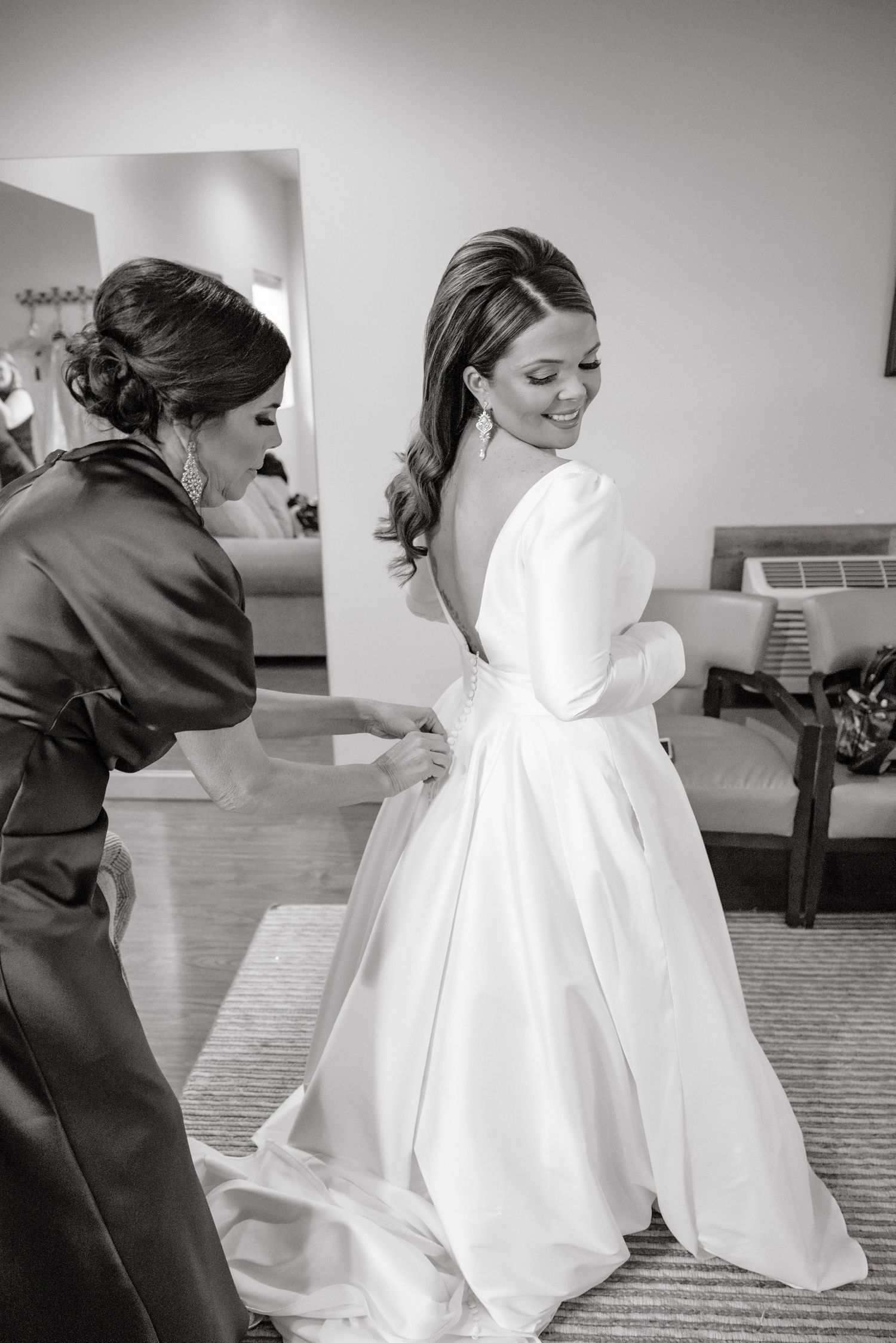Black and white photo of mother helping bride with open back wedding gown before ceremony in bridal suite.