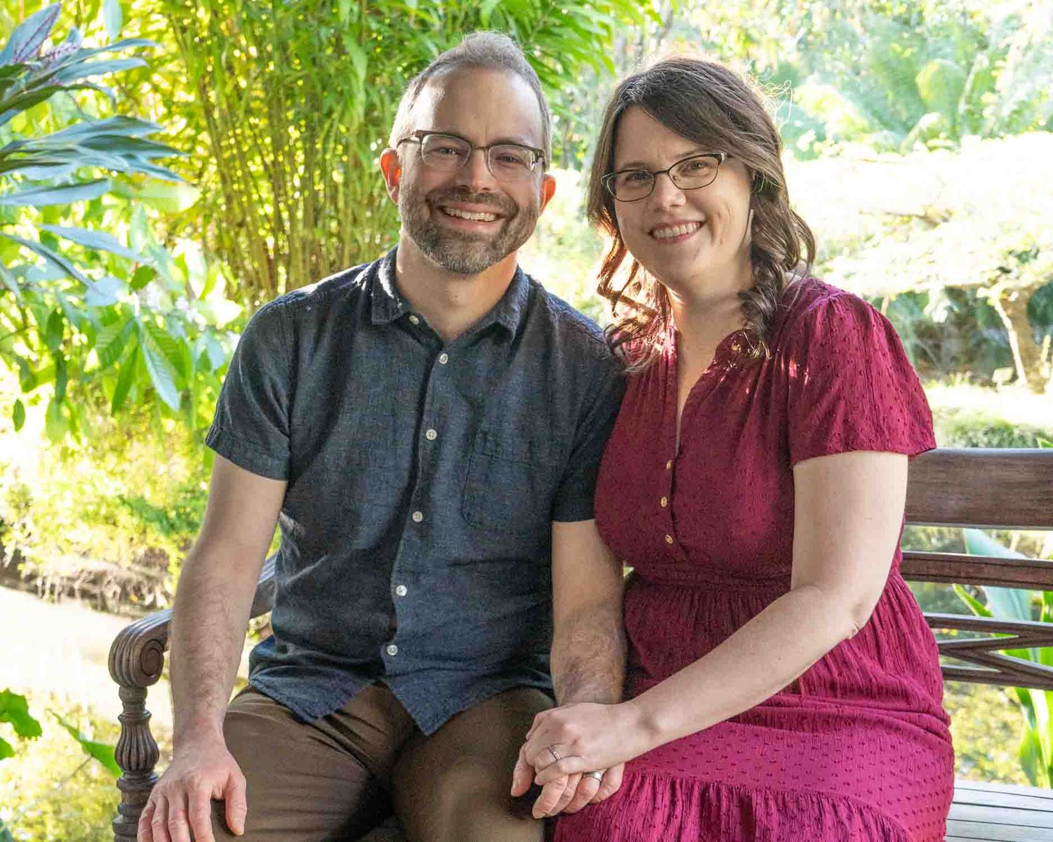 A smiling couple sits together on a park bench surrounded by lush greenery.