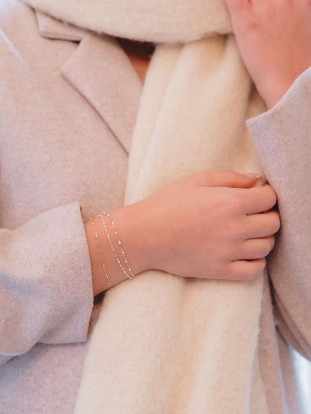 Close-up of a hand wearing delicate gold chain bracelets against a beige coat.