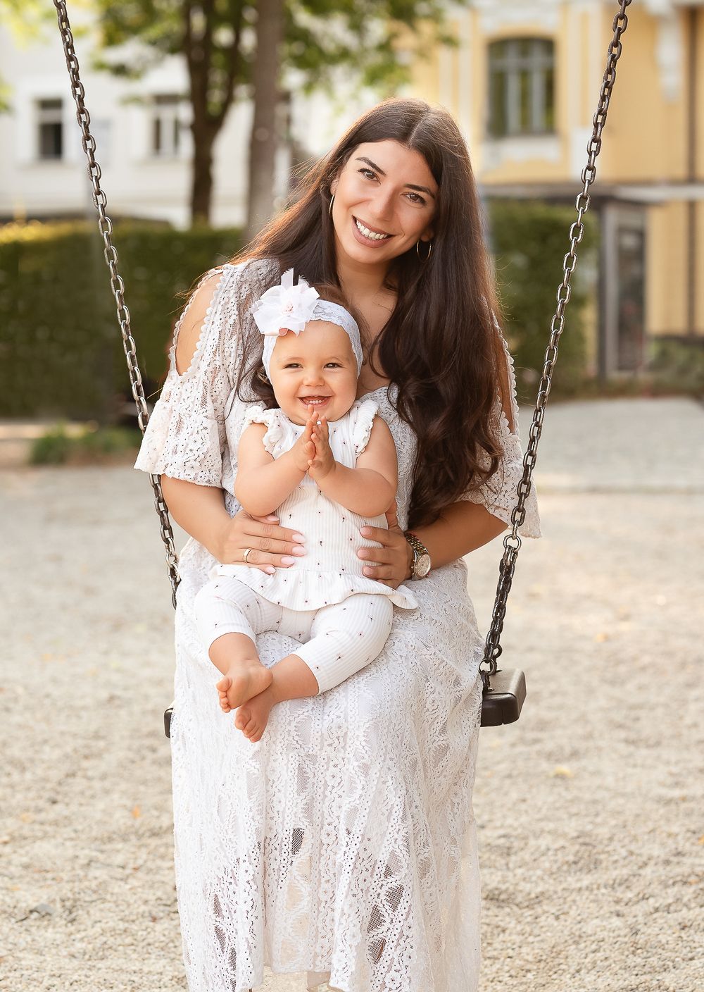 Famillienfotografie - A tender playground moment between a mother and a baby on a swing with white lace dresses and joyful expressions.