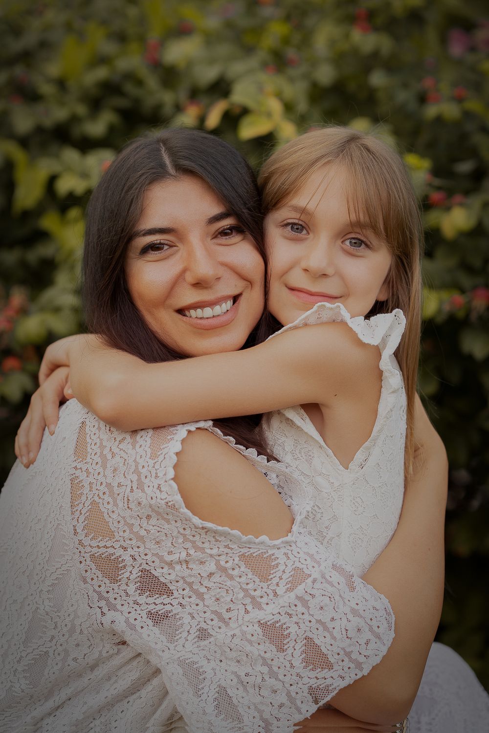 Kinderffotografie - a touching embrace between a mother and a child captured in white lace dresses against a garden backdrop.