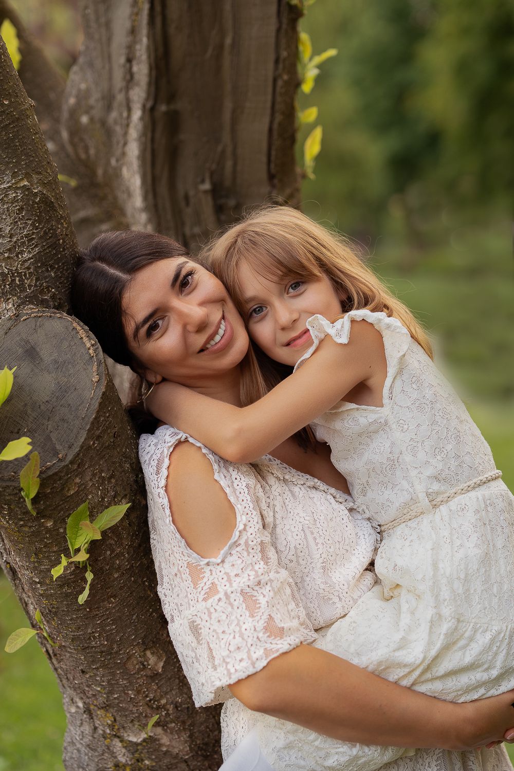 Famillienfotografie - a sweet moment shared between a mother and a child leaning against a tree trunk in matching white lace dresses.
