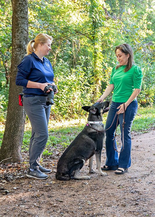 Two people in blue and green shirts stand on a forest trail with a large black German Shepherd dog.
