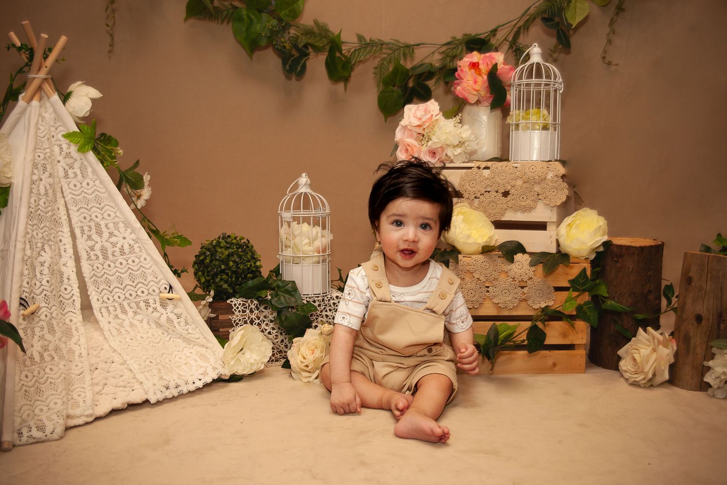 Cute baby sitting on floor beside a white lace teepee with floral decorations and rustic wooden crates.