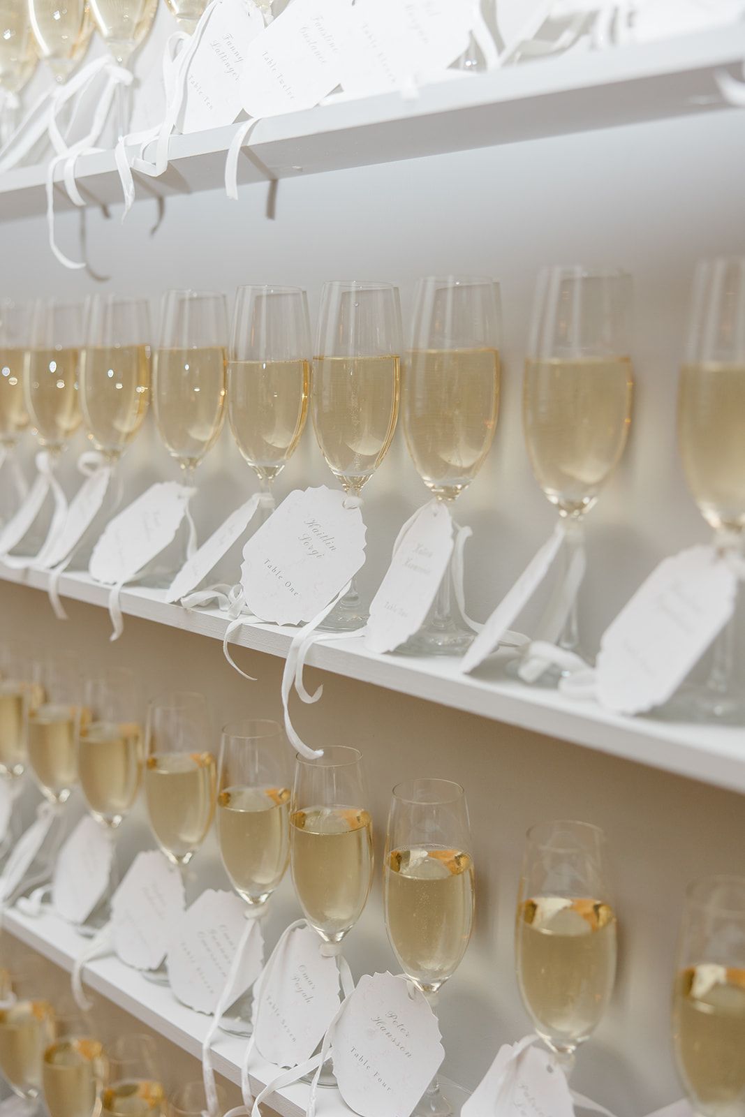 Rows of champagne flutes filled with golden bubbly and decorated with white name tags arranged on elegant display shelves.