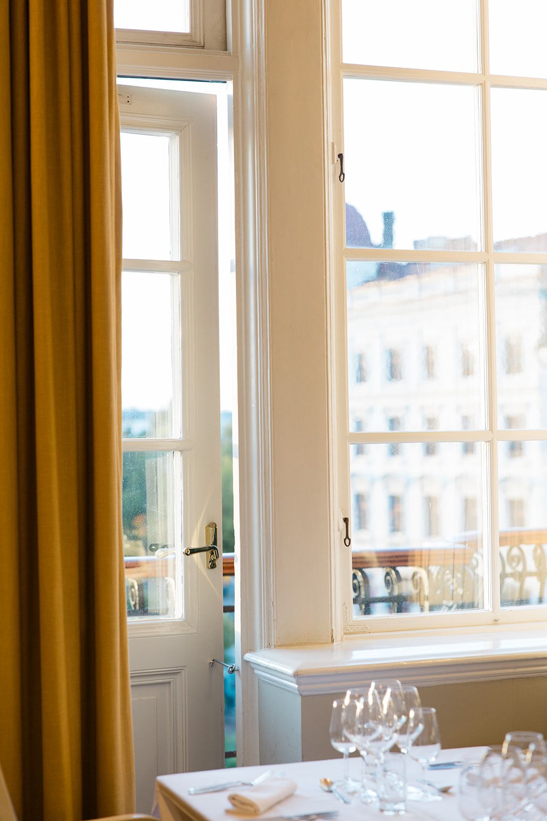 Elegant dining room window with mustard yellow curtains and wine glasses on table overlooking European city buildings.