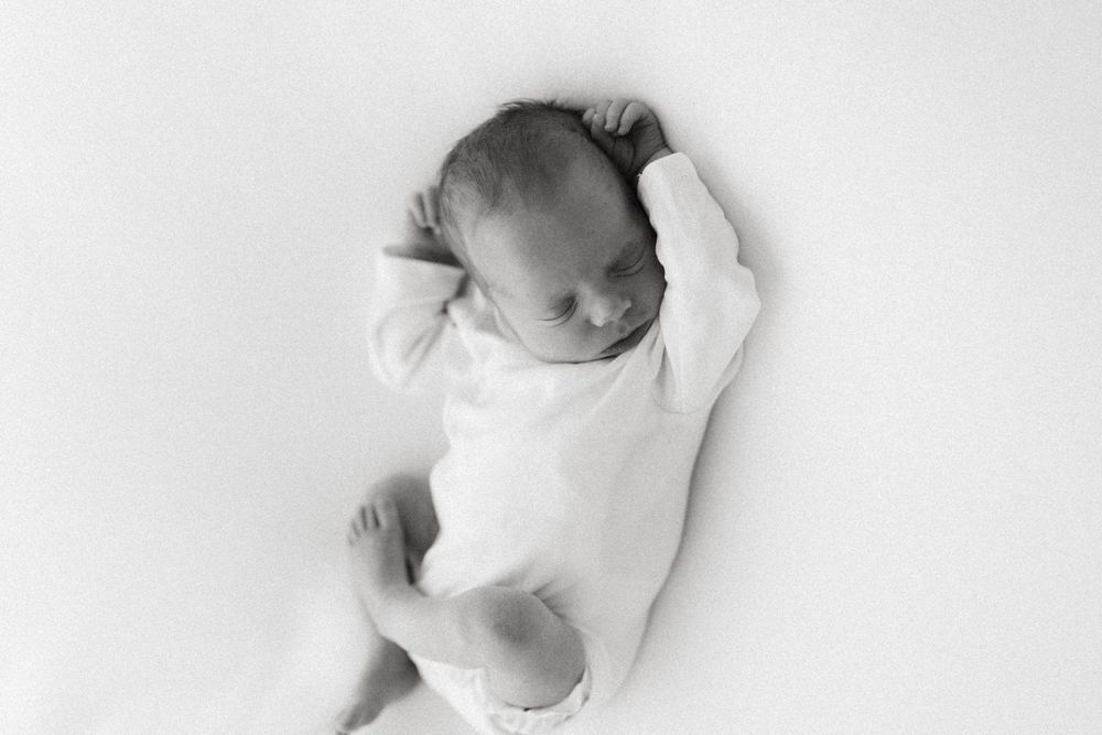 A newborn baby wearing white sleeps peacefully on a white background in black and white photography.