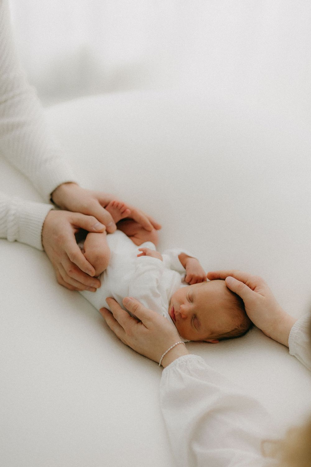 Gentle hands caress a sleeping newborn baby swaddled in soft white fabric against a light background.