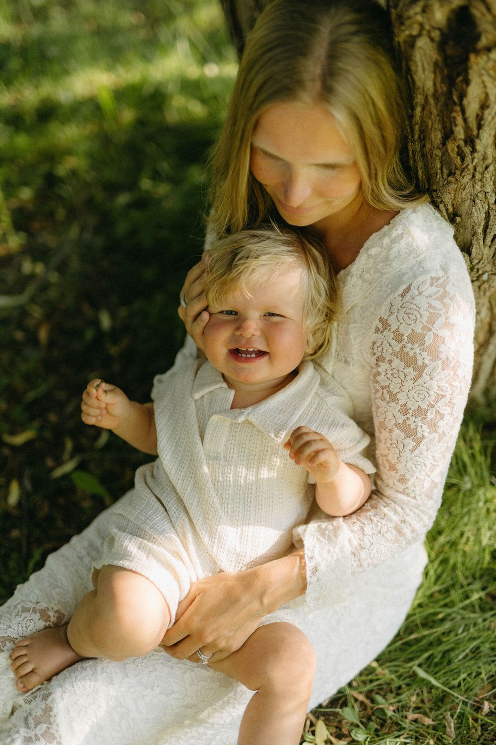 A tender moment captured in white lace dresses as they sit together on grass in dappled sunlight.