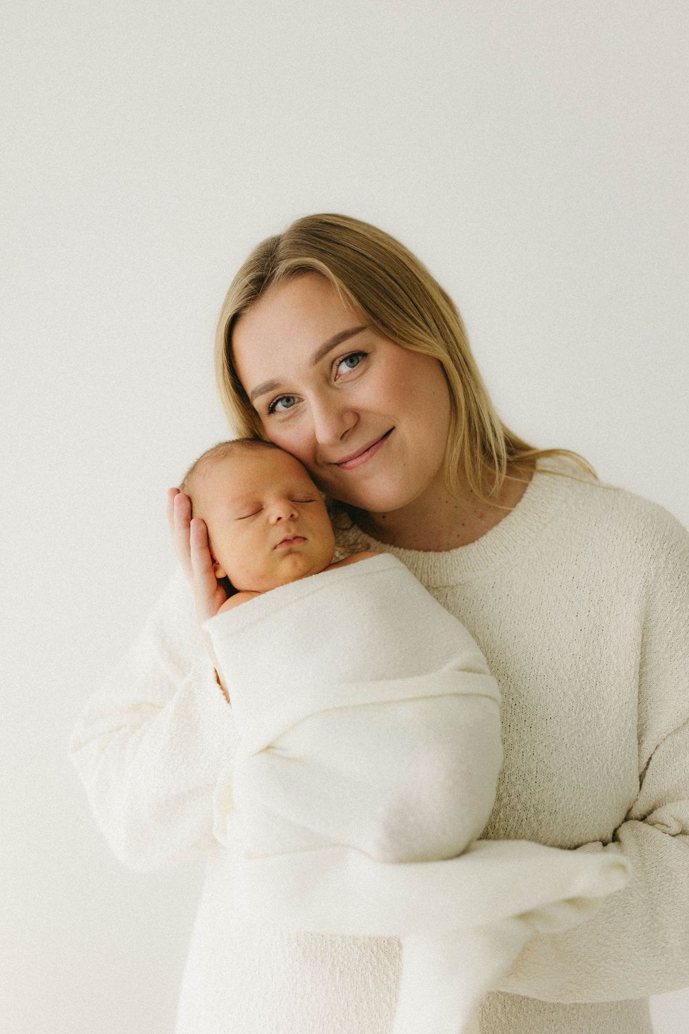 A tender moment captured in soft white tones as a parent holds their sleeping newborn baby swaddled in white fabric.