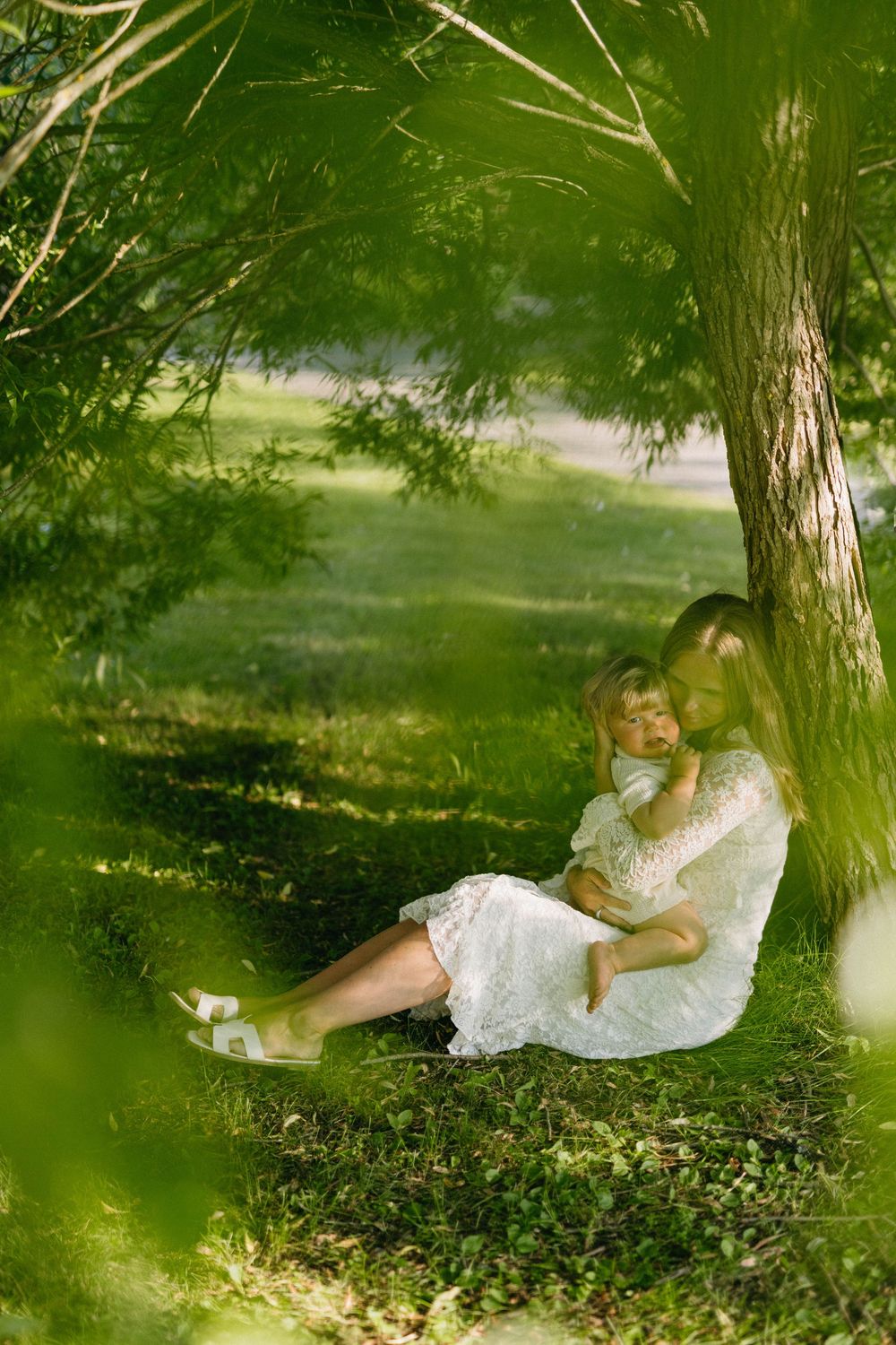 Two figures in white dresses sit together under a shady tree on a sunny summer afternoon in a peaceful garden setting.
