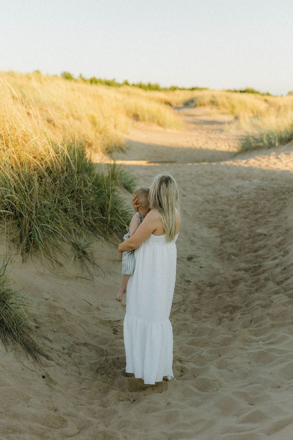 Two figures in white dresses stand together on sandy beach dunes at golden hour.