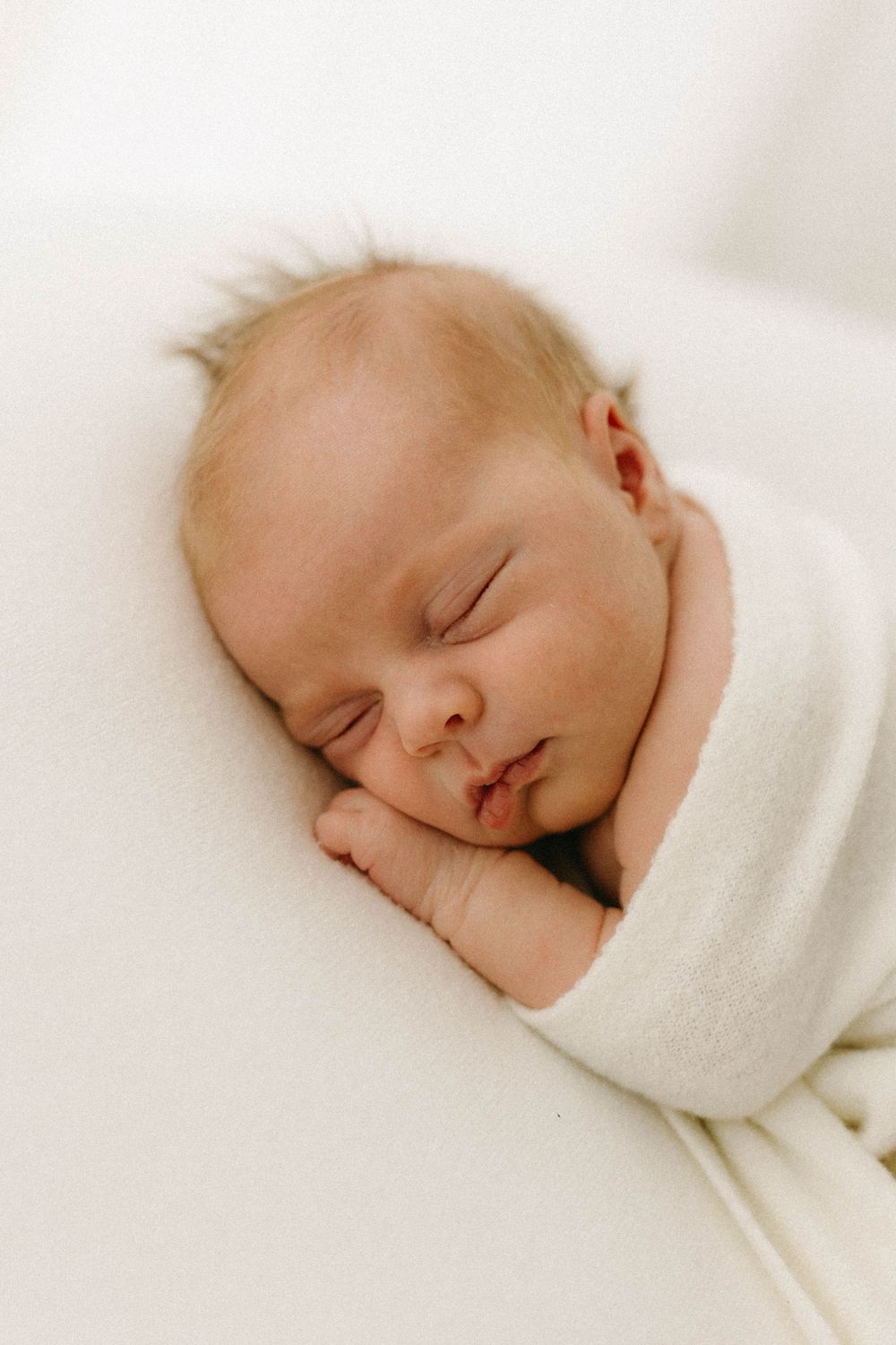 A peaceful sleeping newborn swaddled in soft white blankets rests with closed eyes and gentle expression.