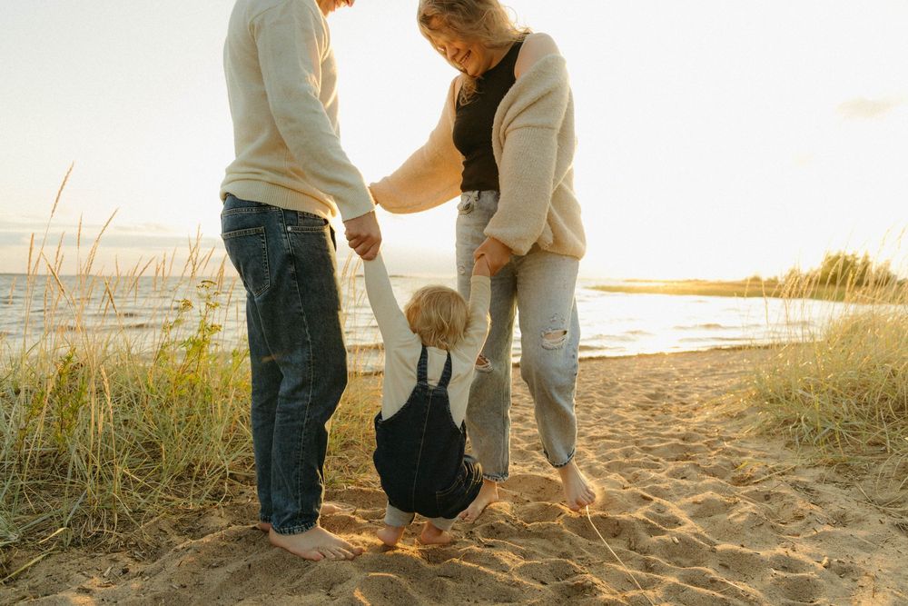 Couple holding hands walking along a sandy beach at sunset with tall grass in the foreground.