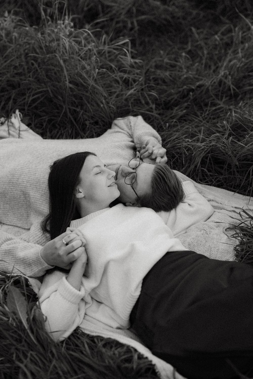 A couple in white sweaters lying together in tall grass on a blanket, captured in black and white photography.