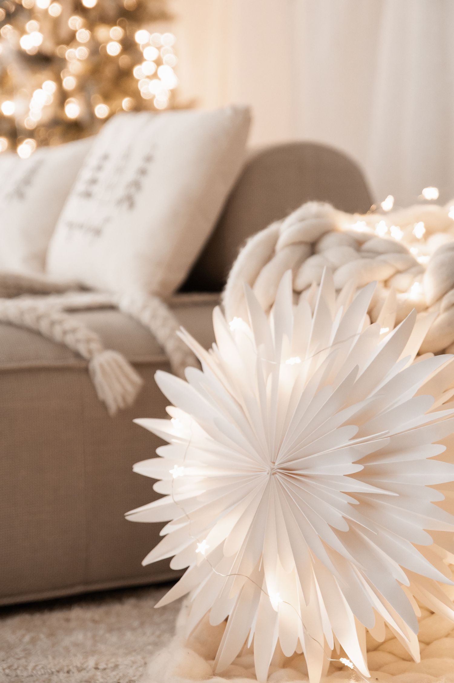 A white paper snowflake decoration sits near cozy pillows with a soft bokeh Christmas tree in the background.