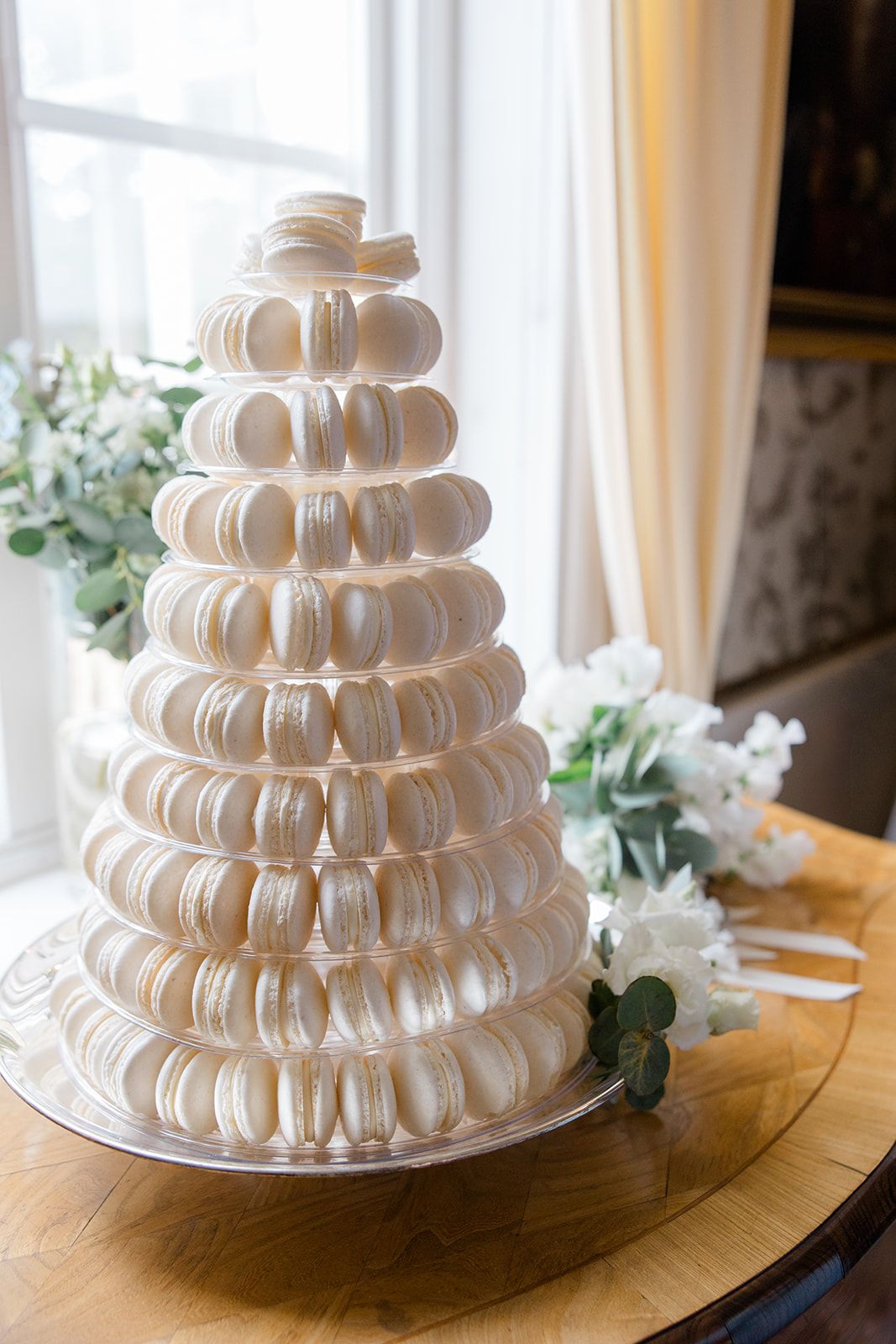 A tiered tower of white macarons arranged in circular layers on a cake stand with eucalyptus greenery accents.