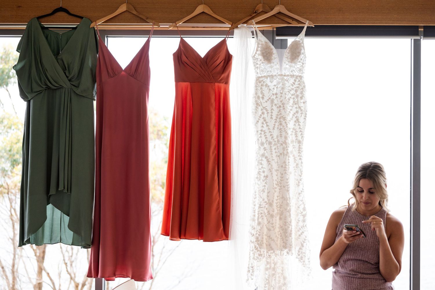 Four formal dresses hanging on display including sage green, burgundy, coral red and white lace gowns by a window.