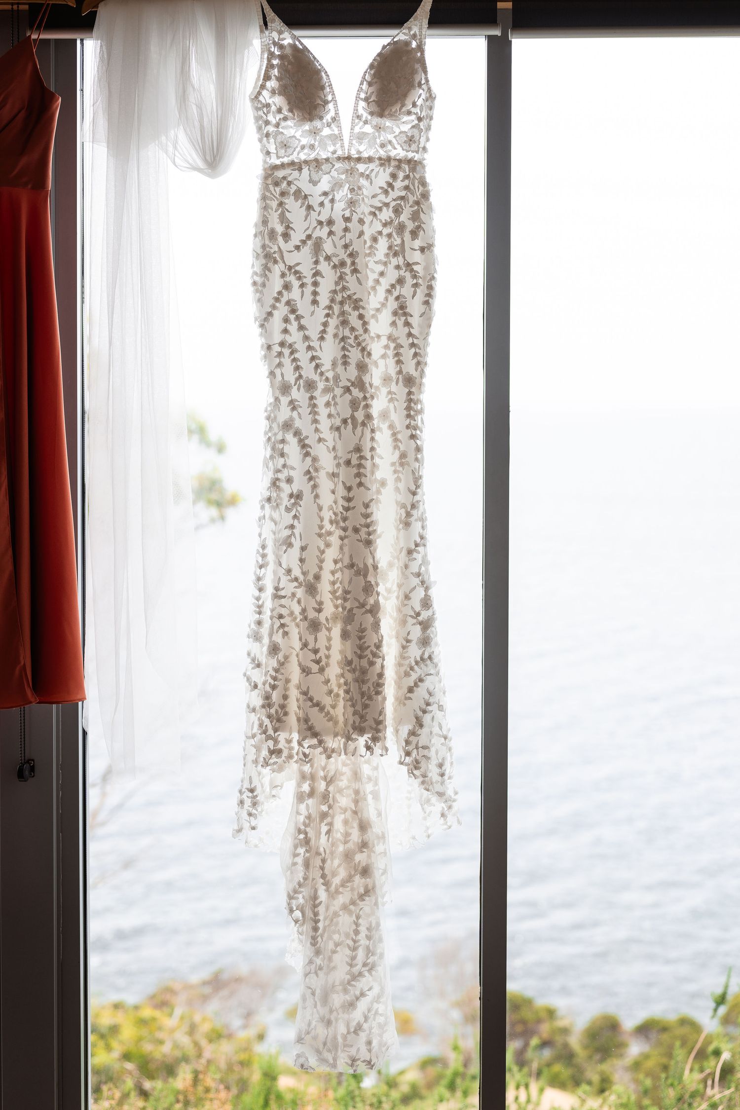 A beaded white wedding dress hangs in front of windows overlooking an ocean view on a sunny day.