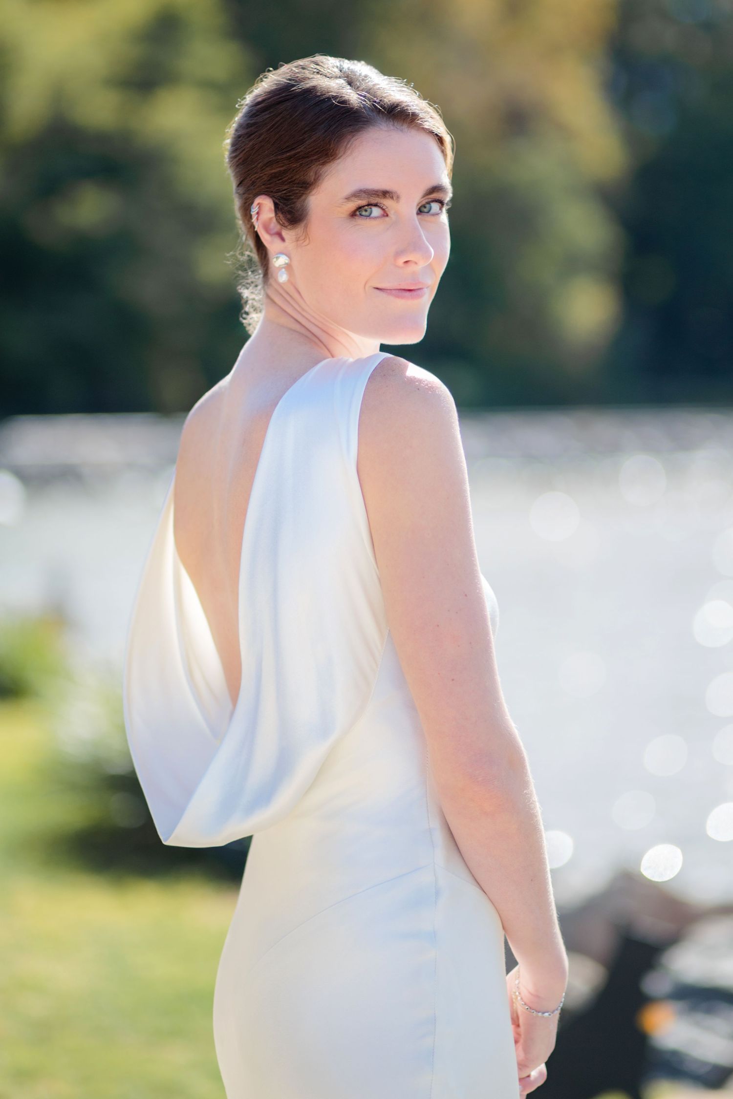 Bride in elegant white sleeveless wedding dress poses outdoors in natural sunlight.