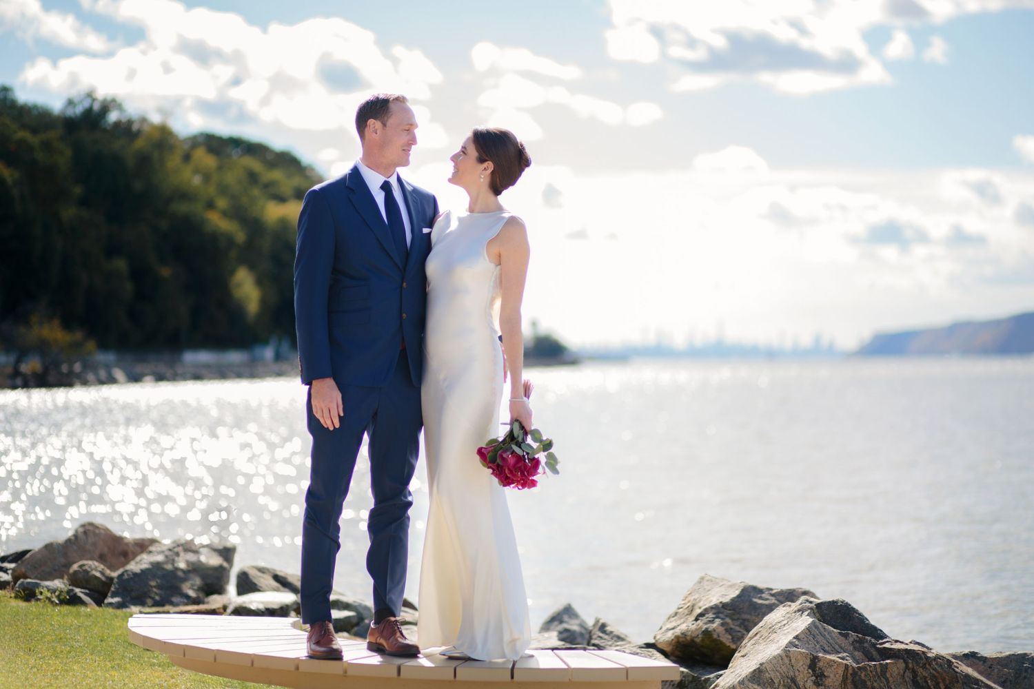A couple in formal wedding attire stands together on a rocky beach shore with mountains, water, and the NYC skyline in the background.