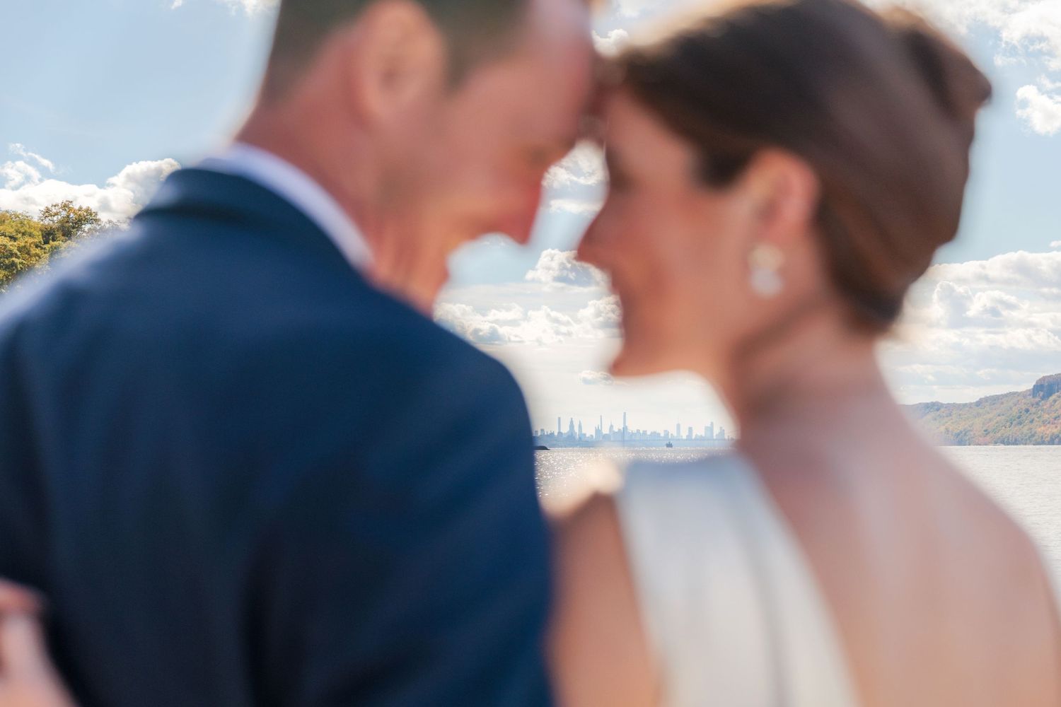A newly wed couple in the forefront of the photo are touching foreheads. They are blurred and between them, the NYC skyline is in focus.