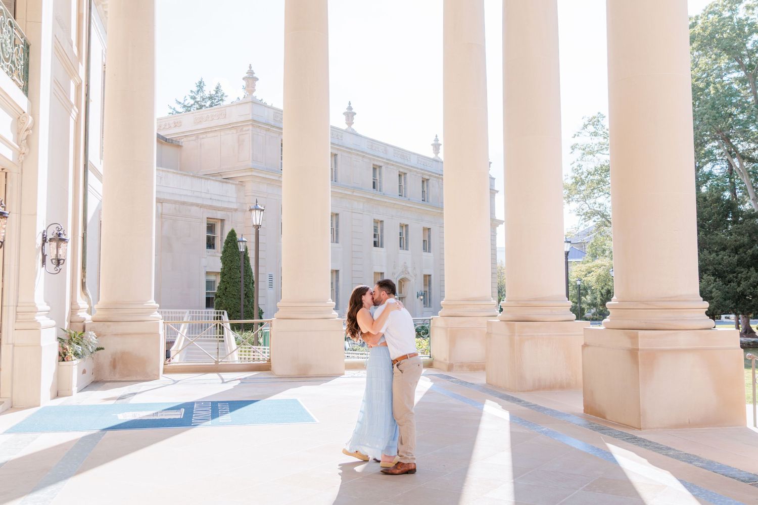 A newly engaged couple embraces on an elegant portico with white columns and a classical mansion in the background.