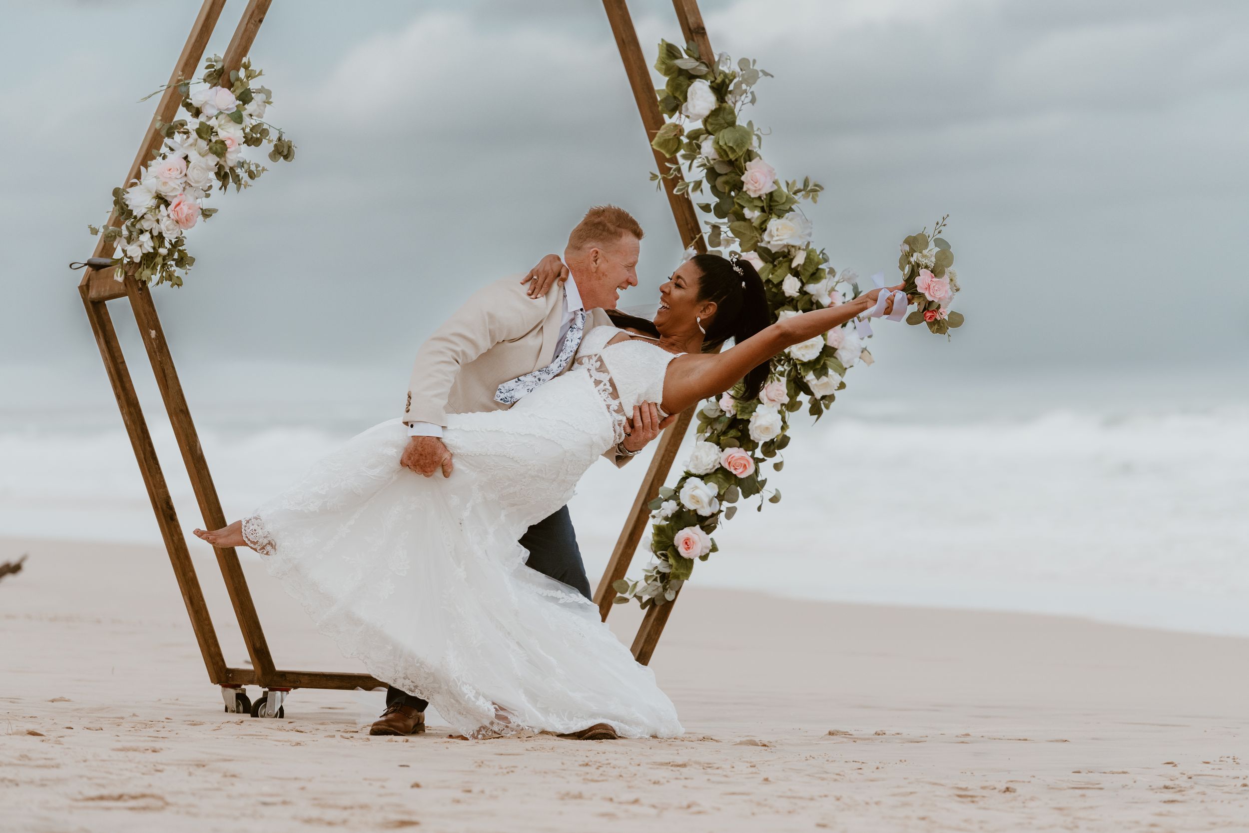 Romantic wedding moment captured on beach under hexagonal floral arch.