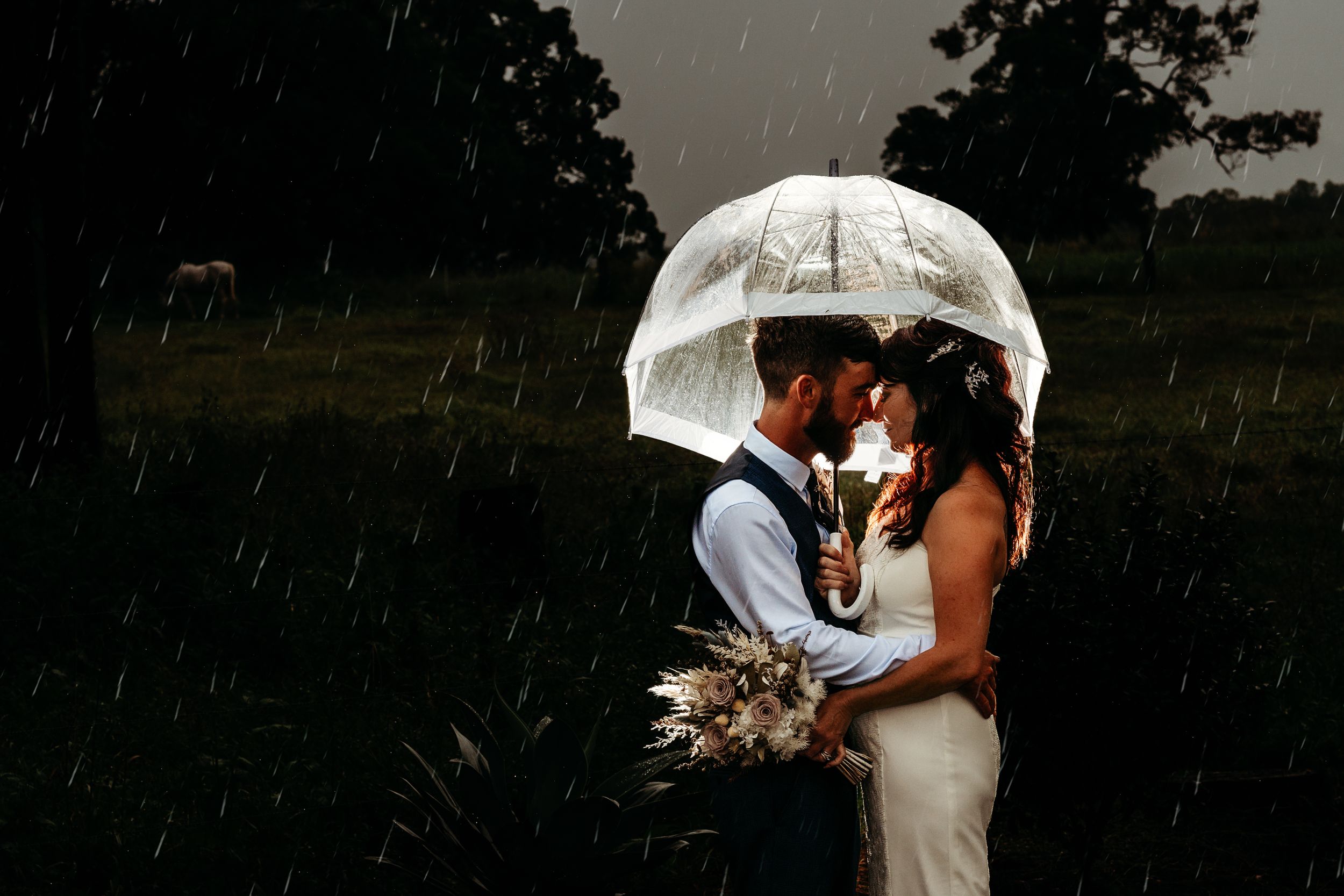 A romantic couple shelters under a clear umbrella in the rain during an outdoor photoshoot.