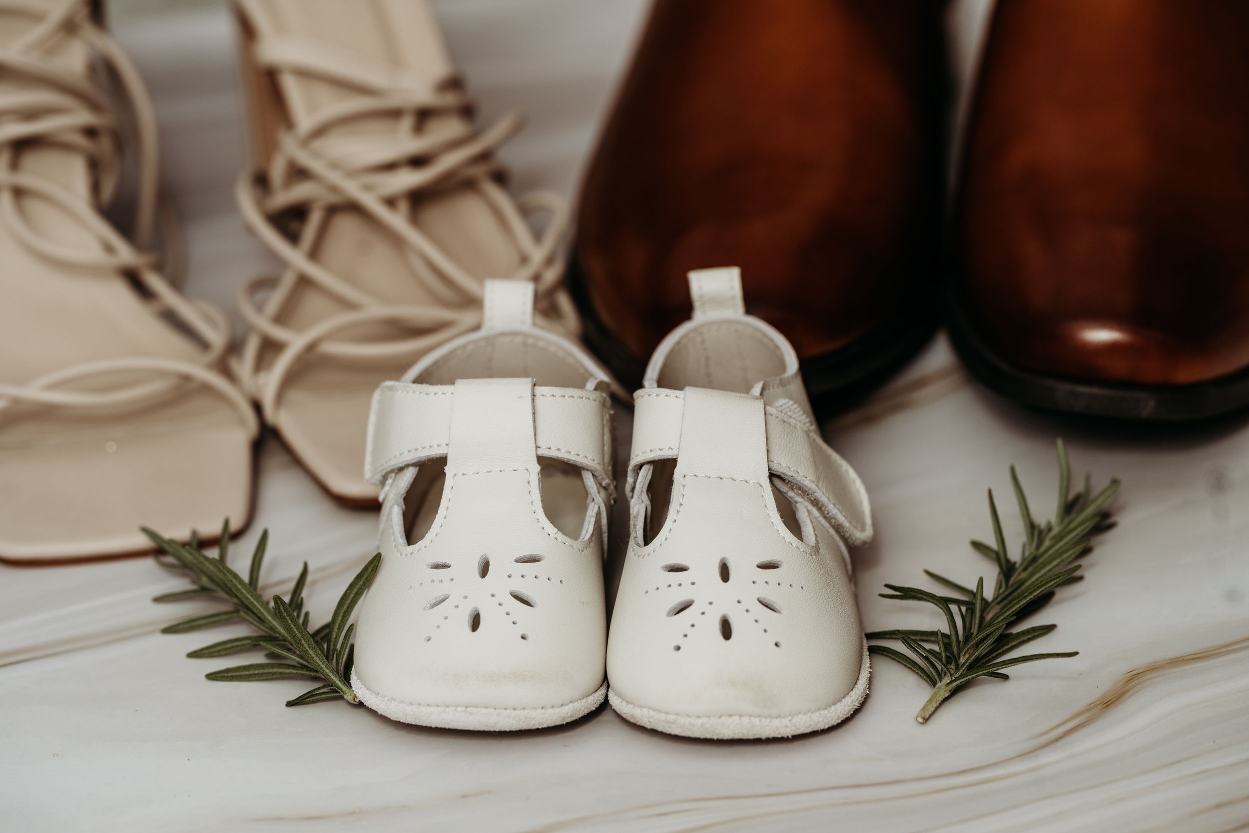 White baby shoes arranged with brown dress shoes and greenery on light background.