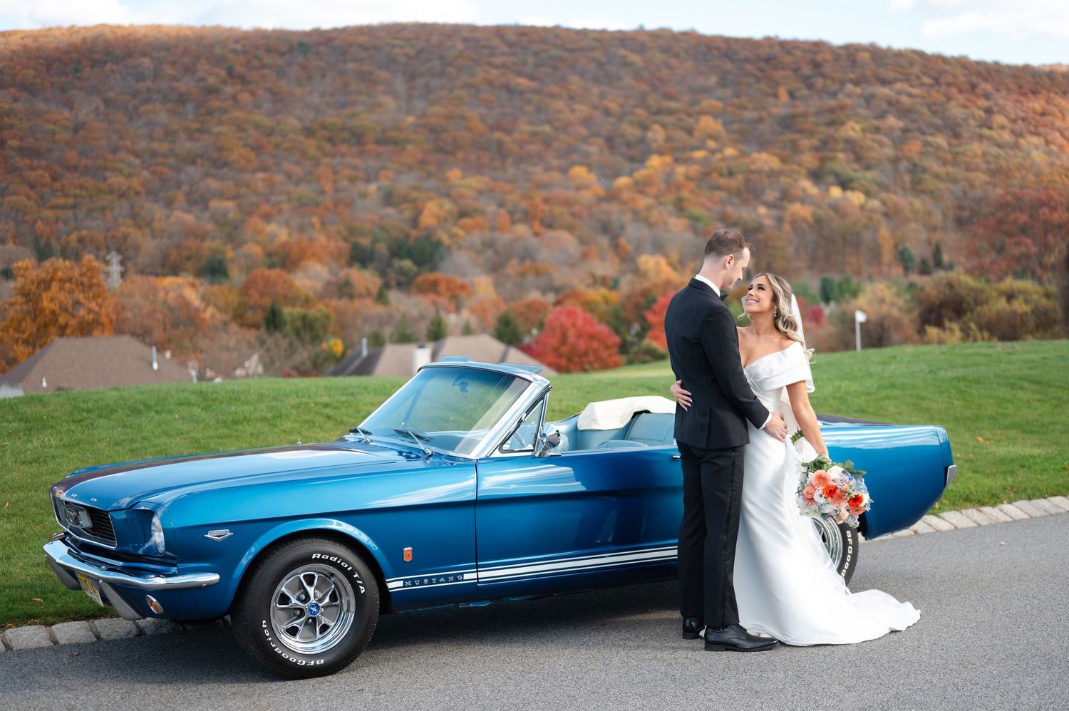 A couple poses for wedding portraits in upstate NY with a vintage blue Mustang surrounded by autumn foliage.