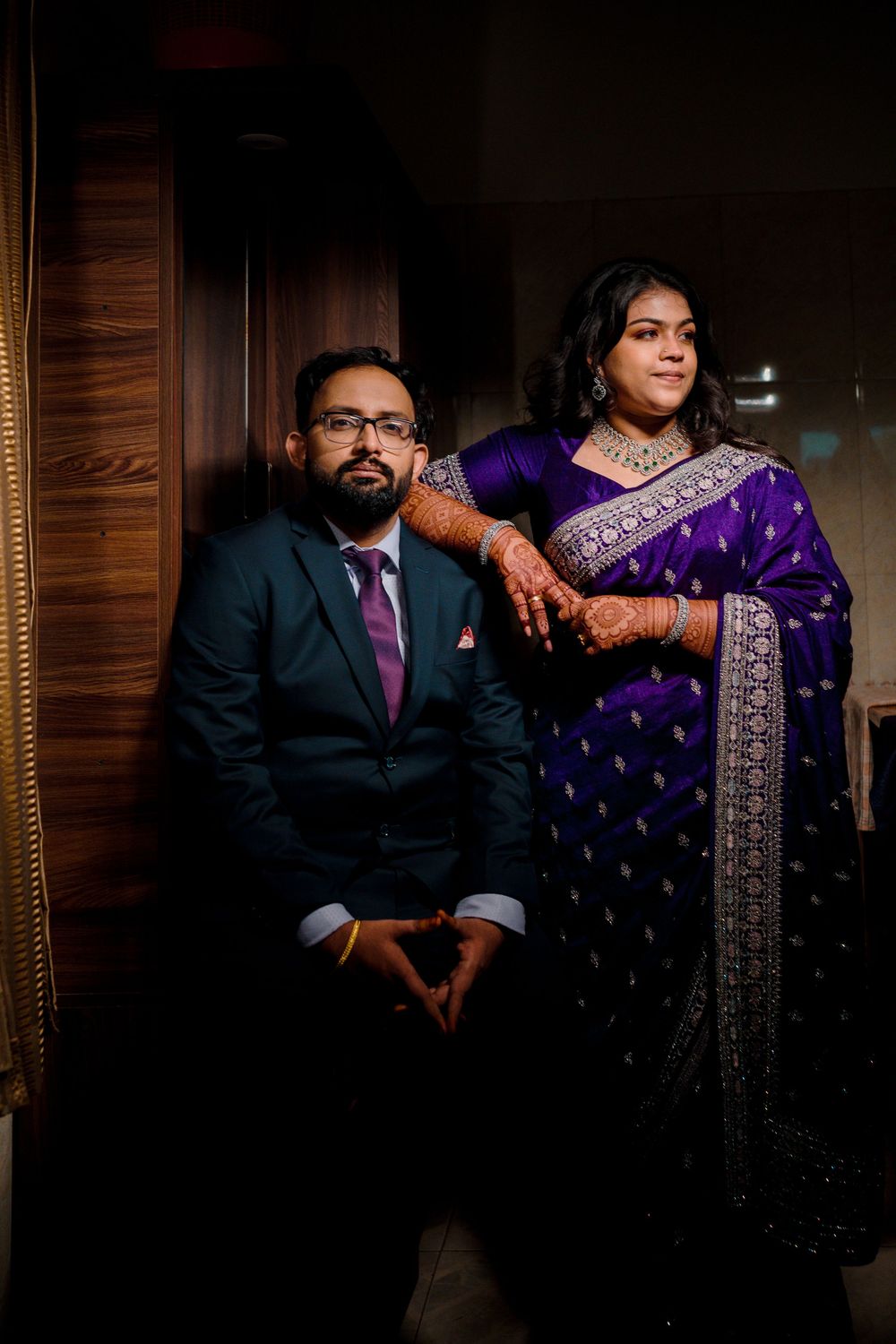 A couple in traditional Indian attire pose together, with the woman wearing a purple sari with silver embellishments.