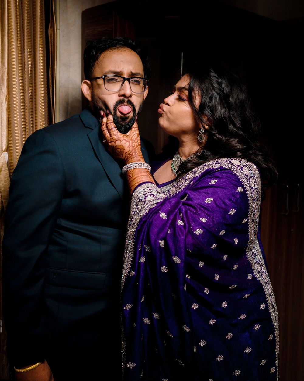 A couple in formal attire share an intimate moment in a dimly lit room with curtains in the background.