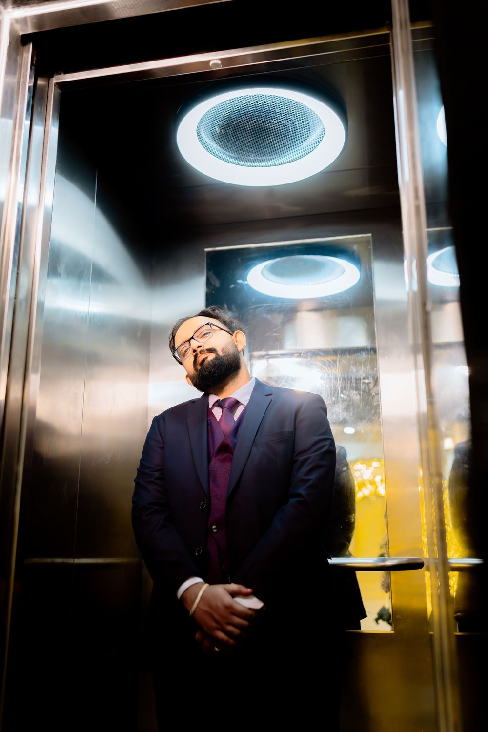 A person in a dark suit and purple tie laughs heartily in a dimly lit elevator with circular overhead lighting.