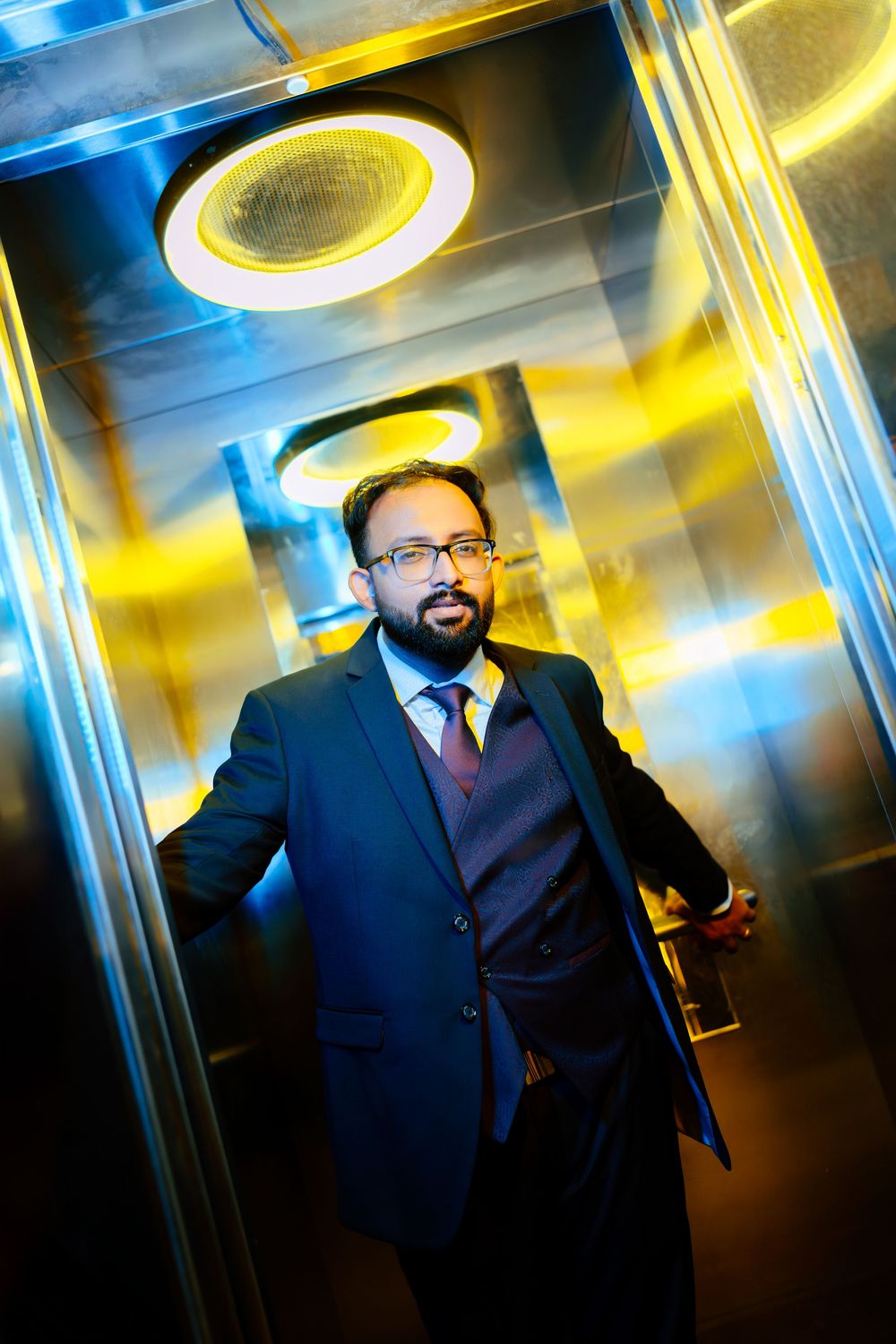Professional portrait in navy suit standing in modern elevator with dramatic blue and yellow lighting.