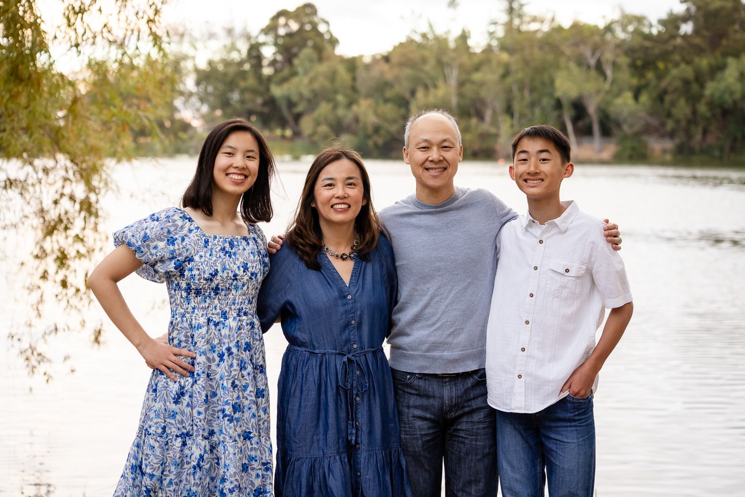 A family wearing coordinated blue and white outfits poses together outdoors by a lake during sunset at Vasona Lake County Park in Los Gatos.