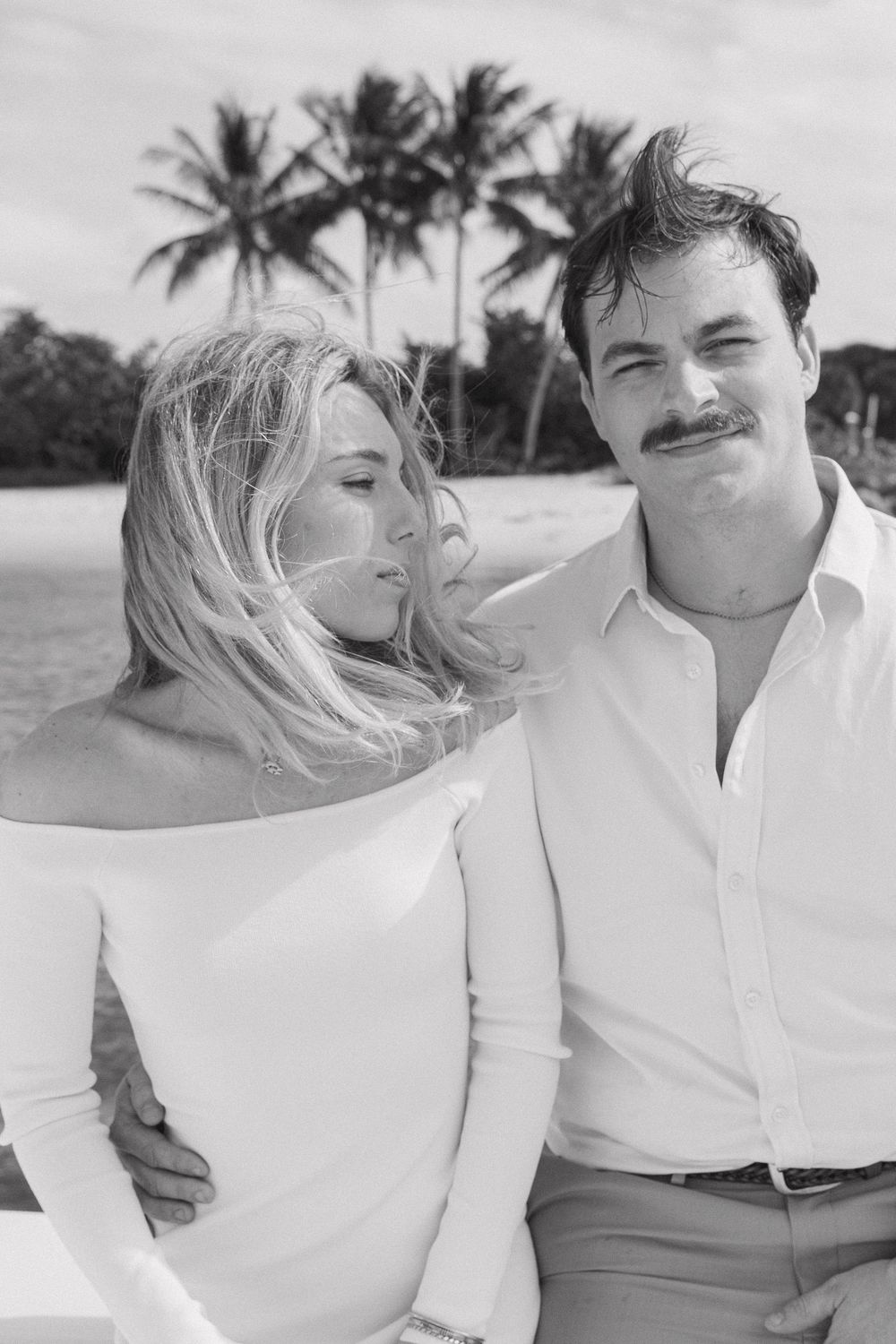 Black and white photo of a couple on a tropical beach with palm trees, woman in off-shoulder dress, man with mustache.