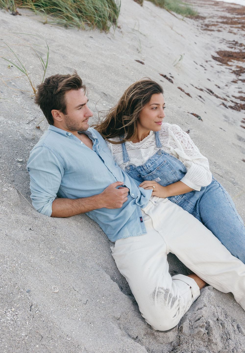Couple relaxing on sandy beach dunes, man in blue shirt and woman in denim overalls enjoying a serene coastal view.