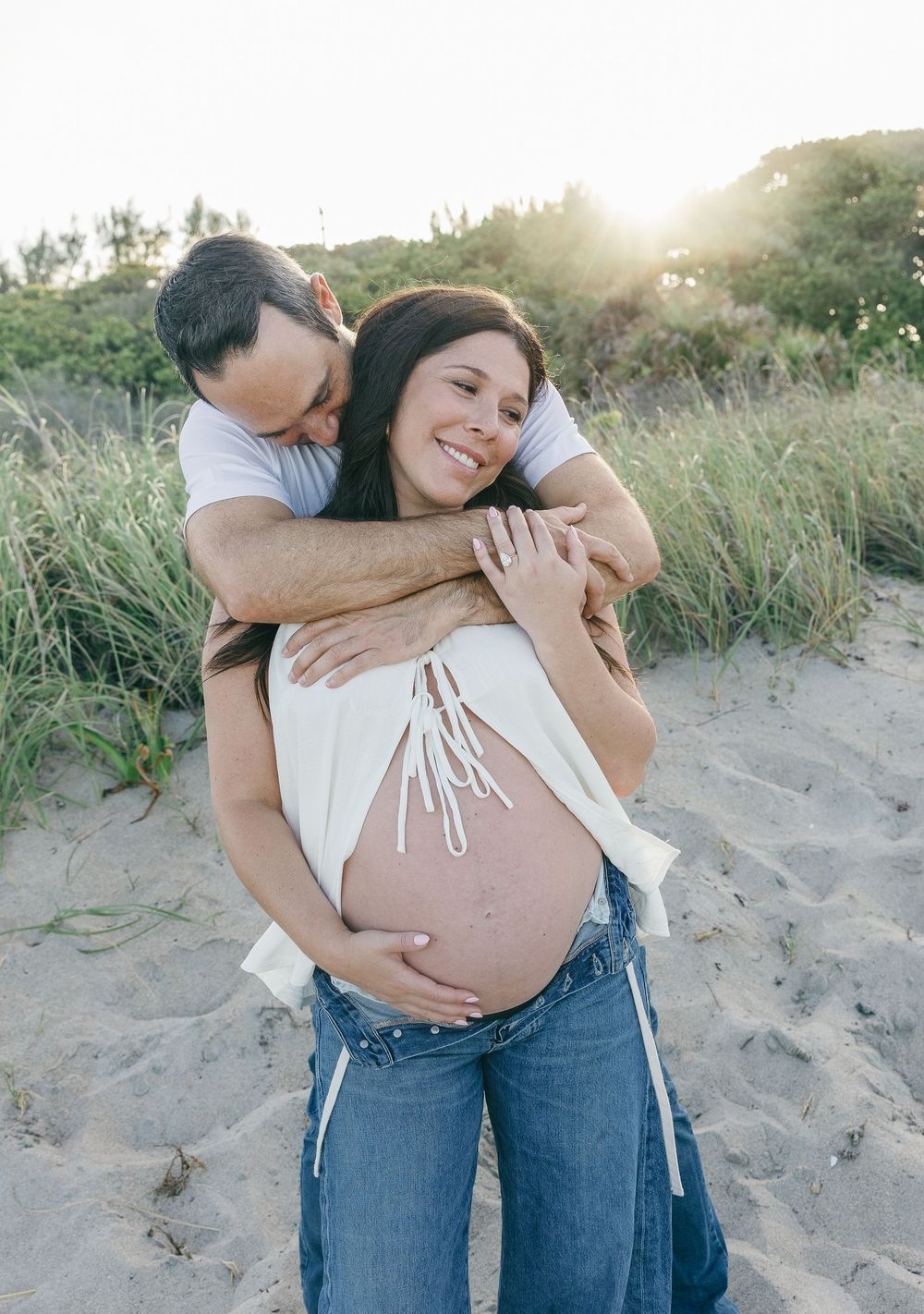 A couple shares a tender embrace on a sandy beach, with the man hugging his pregnant partner from behind at sunset.
