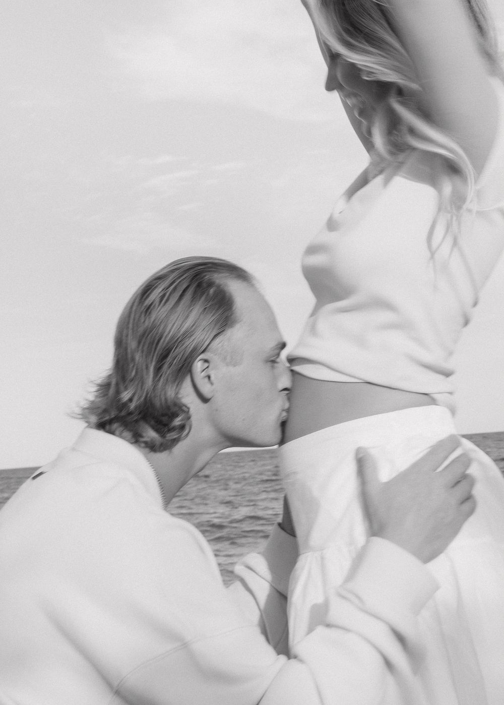 Black and white photo of a man kissing a pregnant woman's bare belly by the ocean.