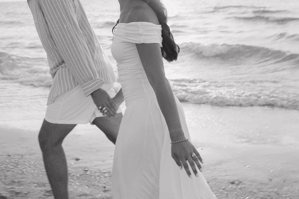 A couple in white outfits walking along a sandy beach at sunset holding hands and embracing.