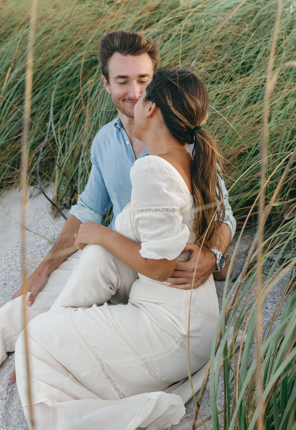 Couple sitting in beach grass, woman in white dress embraced by man in blue shirt during romantic outdoor engagement shoot.