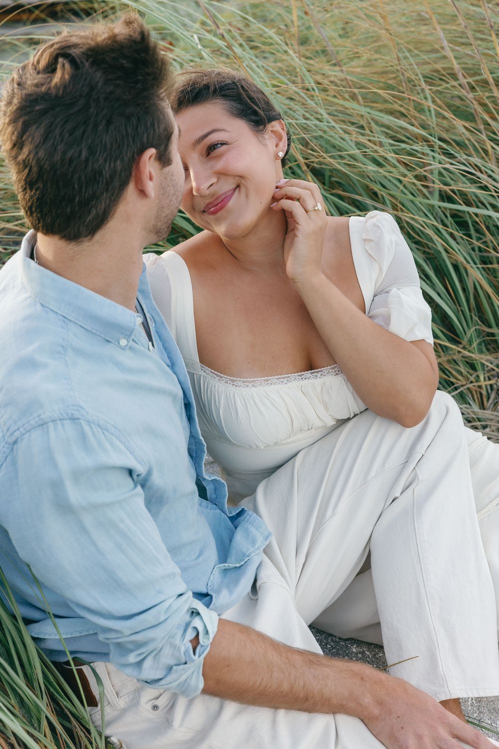 Happy couple sitting together in beach grass, woman smiling while touching her face, wearing white outfit.