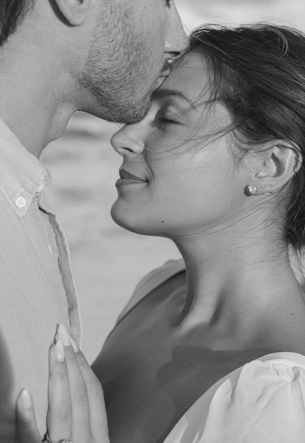 Black and white close-up of a romantic couple almost kissing, woman smiling with pearl earrings outdoors.